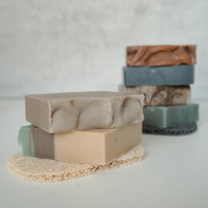 May include: A stack of four handmade soap bars in various shades of brown, gray, and green, resting on a natural loofah sponge. The soap bars are arranged in a pyramid shape, with the largest bar at the bottom and the smallest bar at the top.
