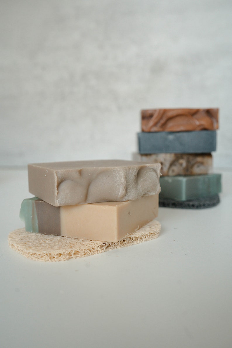 May include: A stack of four handmade soap bars in various shades of brown, gray, and green, resting on a natural loofah sponge. The soap bars are arranged in a pyramid shape, with the largest bar at the bottom and the smallest bar at the top.