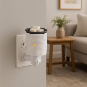 May include: A white plug-in wax warmer with a black rim and a decorative arched pattern. The warmer contains three white wax cubes and an illuminated orange light. The warmer is plugged into a white wall outlet.