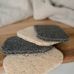 May include: Three loofah soap pads, two are dark gray and one is light beige. The pads are stacked on top of each other on a wooden surface.