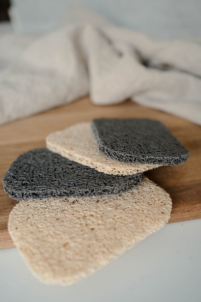 May include: Three loofah soap pads, two are dark gray and one is light beige. The pads are stacked on top of each other on a wooden surface.