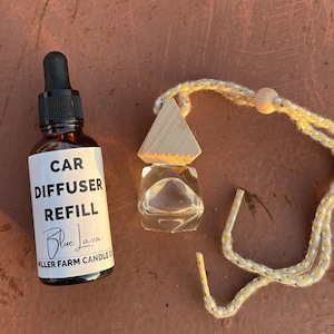 May include: A car air freshener refill bottle and a car diffuser. The brown glass bottle has a dropper and a label that reads "CAR DIFFUSER REFILL Blue Java". The car diffuser is clear with a wooden top and a rope for hanging.