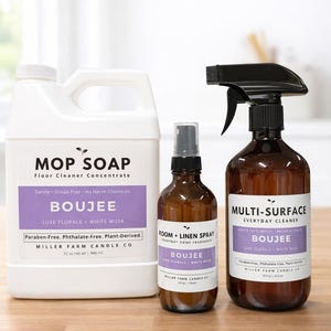 May include: A collection of cleaning products from Miller Farm Candle Co. includes a white jug of floor cleaner, a brown spray bottle of multi-surface cleaner, and a brown room and linen spray. All products feature the "Boujee" scent.