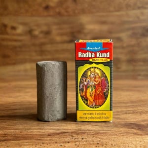 May include: A dark gray, cylindrical tilak stick next to a box of Radha Kund Dark Tilak. The box has a red top and a yellow bottom with an illustration of two figures. Text on the box includes "Scented Radha Kund DARK TILAK".