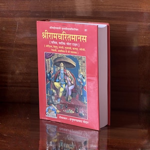 May include: A red hardcover book with text in a foreign language and a colourful illustration. The title "Shriramcharitmanas" is displayed. The book's spine is visible, and the pages are white.