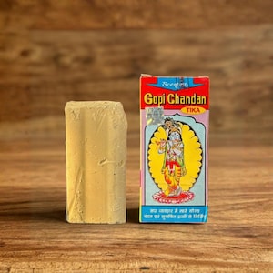 Può includere: Un blocco rettangolare beige chiaro di Gopi Chandan Tika accanto a una scatola colorata. La scatola presenta un'illustrazione di una divinità con un alone giallo e testo in più lingue. Lo sfondo è una superficie di legno.