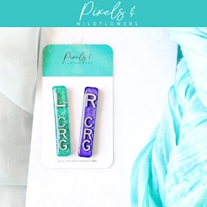 May include: Two resin hair clips, one teal and one purple, each with silver letters spelling "LCRG" and "RCRG". The clips are displayed on a white card with the text "Pixels & Wildflowers" and "Let all that you do be done in love."