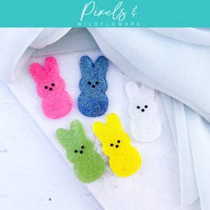 May include: Five colorful bunny-shaped resin embellishments with glitter. The bunnies are pink, blue, white, green, and yellow.