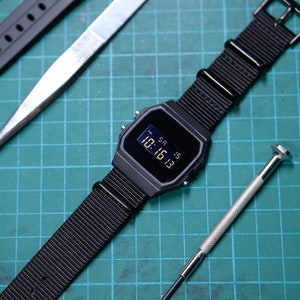 May include: A black digital watch with a black nylon strap, a black rubber strap, a silver screwdriver, and a pair of silver tweezers on a green cutting mat.