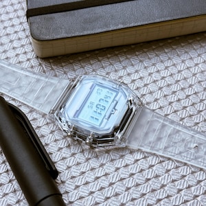 Puede incluir: Un reloj digital transparente con una esfera rectangular y una correa transparente. El reloj muestra la hora y la fecha. Un bol&iacute;grafo negro y un cuaderno negro tambi&eacute;n est&aacute;n en la imagen.