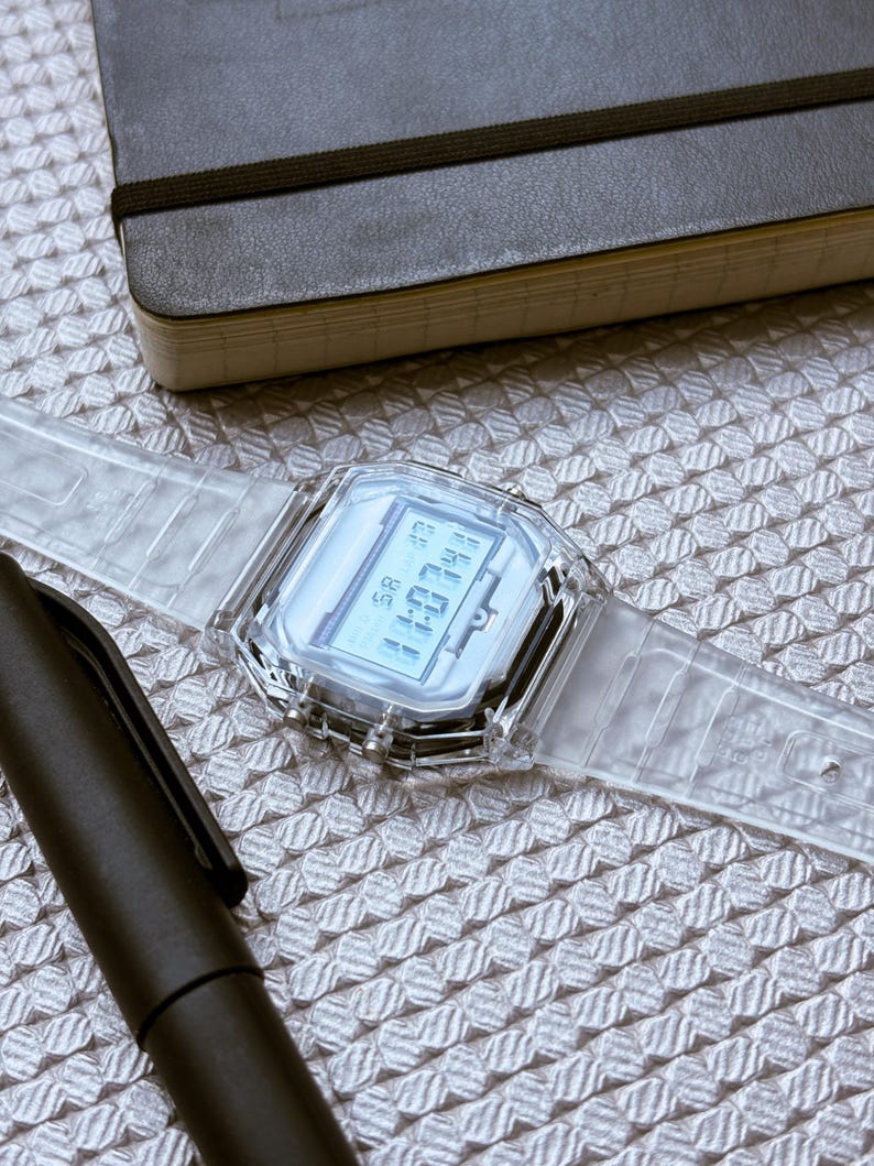 Puede incluir: Un reloj digital transparente con una esfera rectangular y una correa transparente. El reloj muestra la hora y la fecha. Un bol&iacute;grafo negro y un cuaderno negro tambi&eacute;n est&aacute;n en la imagen.