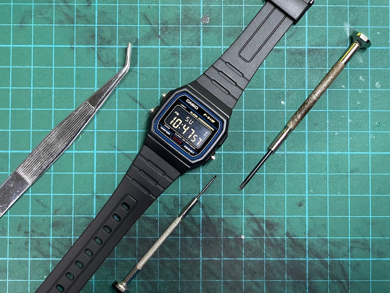 Peut inclure: Une montre num&eacute;rique noire Casio F-91W avec un bracelet en r&eacute;sine noire et un &eacute;cran num&eacute;rique bleu affichant l'heure 10:47:51. La montre est sur un tapis de coupe vert avec deux tournevis et une pince &agrave; &eacute;piler.