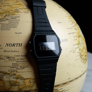 Op de afbeelding: Een zwart digitaal horloge met een zwarte resin band is geplaatst op een globe. Het horloge gezicht toont de tijd 11:35. De globe is in vintage stijl met een beige achtergrond en bruine lijnen.