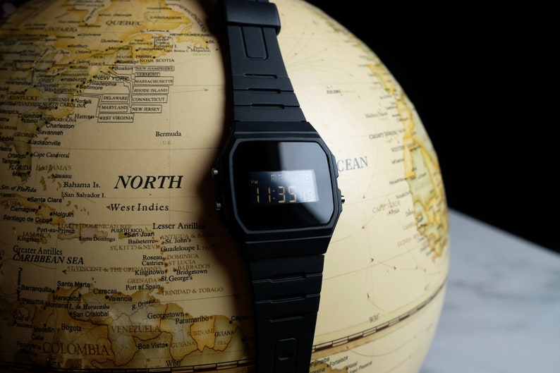 Op de afbeelding: Een zwart digitaal horloge met een zwarte resin band is geplaatst op een globe. Het horloge gezicht toont de tijd 11:35. De globe is in vintage stijl met een beige achtergrond en bruine lijnen.
