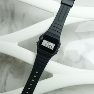 Op de afbeelding: Een zwart digitaal polshorloge met een zwarte band. Het horloge laat de tijd 7:14:33 zien en de dag van de week, "WE", wat staat voor woensdag.