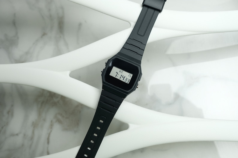 Op de afbeelding: Een zwart digitaal polshorloge met een zwarte band. Het horloge laat de tijd 7:14:33 zien en de dag van de week, "WE", wat staat voor woensdag.