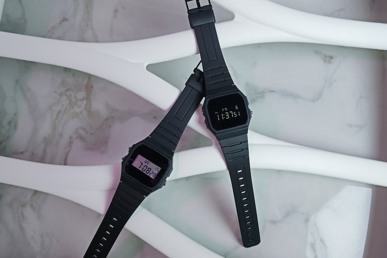 Op de afbeelding: Twee zwarte digitale polshorloges met zwarte banden op een witte marmeren achtergrond. Het bovenste horloge toont de tijd "1:13:51" en het onderste horloge toont de tijd "1:08:29".