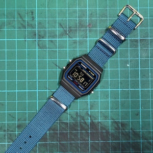 Peut inclure: Une montre num&eacute;rique noire Casio F-91W avec un bracelet en nylon bleu. Le cadran de la montre affiche l'heure, la date et le jour de la semaine. La montre est sur un tapis de coupe vert.