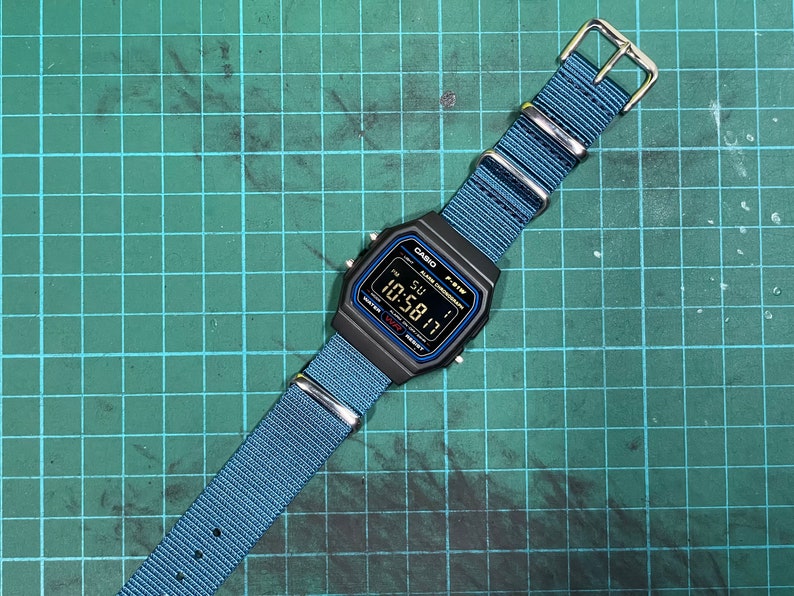 Peut inclure: Une montre num&eacute;rique noire Casio F-91W avec un bracelet en nylon bleu. Le cadran de la montre affiche l'heure, la date et le jour de la semaine. La montre est sur un tapis de coupe vert.