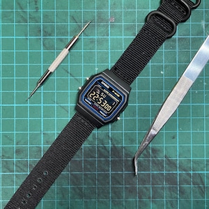 Peut inclure: Une montre num&eacute;rique noire avec un bracelet en nylon noir, un outil argent&eacute; et une &eacute;pingle argent&eacute;e sur un tapis de coupe vert. Le cadran de la montre affiche l'heure 22:53:00.