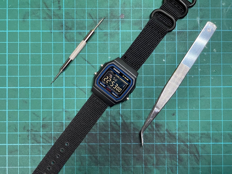 Peut inclure: Une montre num&eacute;rique noire avec un bracelet en nylon noir, un outil argent&eacute; et une &eacute;pingle argent&eacute;e sur un tapis de coupe vert. Le cadran de la montre affiche l'heure 22:53:00.