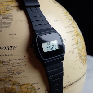 Op de afbeelding: Een zwart digitaal polshorloge met een zwarte harsband ligt op een globe. Het horloge laat de tijd 7:022 en de dag van de week, WE, zien.