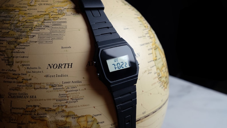 Op de afbeelding: Een zwart digitaal polshorloge met een zwarte harsband ligt op een globe. Het horloge laat de tijd 7:022 en de dag van de week, WE, zien.