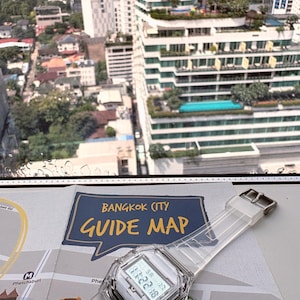 Puede incluir: Un reloj digital transparente con esfera blanca y correa transparente descansa sobre un mapa con la leyenda "Bangkok City Guide Map". El reloj muestra la fecha y la hora. El fondo muestra un paisaje urbano con edificios altos.