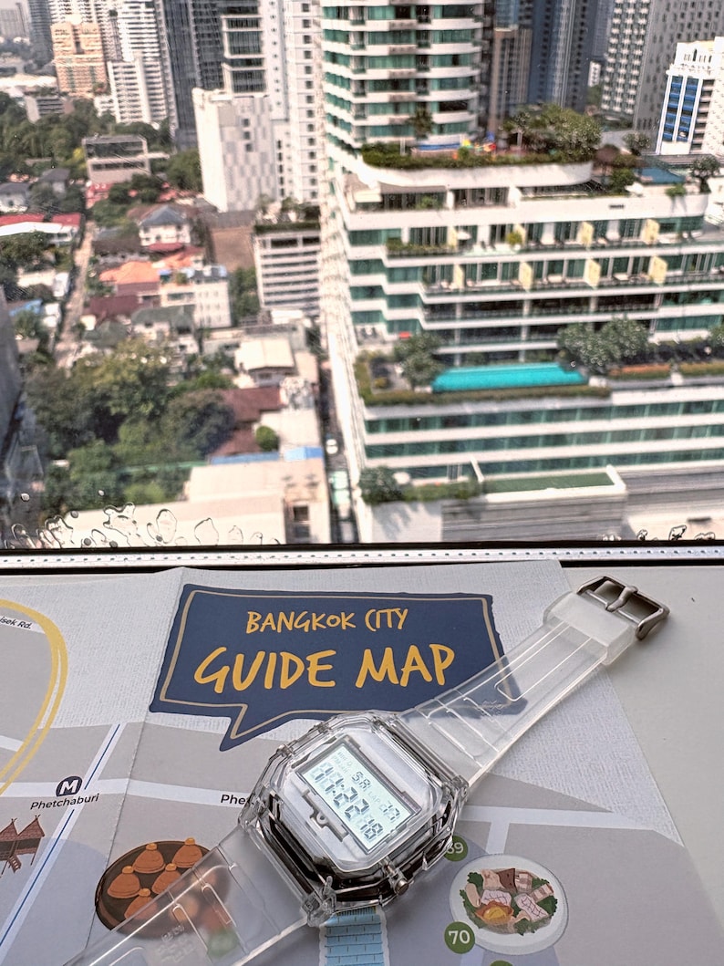 Puede incluir: Un reloj digital transparente con esfera blanca y correa transparente descansa sobre un mapa con la leyenda "Bangkok City Guide Map". El reloj muestra la fecha y la hora. El fondo muestra un paisaje urbano con edificios altos.
