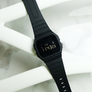Op de afbeelding: Een zwart digitaal polshorloge met een zwarte band. Het horloge laat de tijd 11:43:23 zien en de dag FR 6.