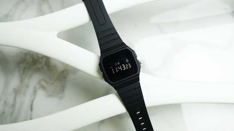 Op de afbeelding: Een zwart digitaal polshorloge met een zwarte band. Het horloge laat de tijd 11:43:23 zien en de dag FR 6.