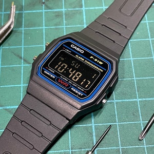 Peut inclure: Une montre num&eacute;rique noire Casio F-91W avec une bordure bleue autour du cadran. Le cadran de la montre affiche l'heure, la date et le jour de la semaine. La montre est sur un bracelet en r&eacute;sine noire.