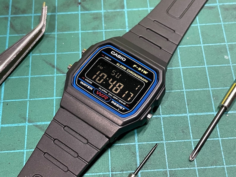 Peut inclure: Une montre num&eacute;rique noire Casio F-91W avec une bordure bleue autour du cadran. Le cadran de la montre affiche l'heure, la date et le jour de la semaine. La montre est sur un bracelet en r&eacute;sine noire.