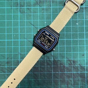 Peut inclure: Une montre num&eacute;rique noire Casio F-91W avec un bracelet en nylon beige. Le cadran de la montre affiche l'heure, la date et le jour de la semaine. La montre est sur un tapis de coupe vert.
