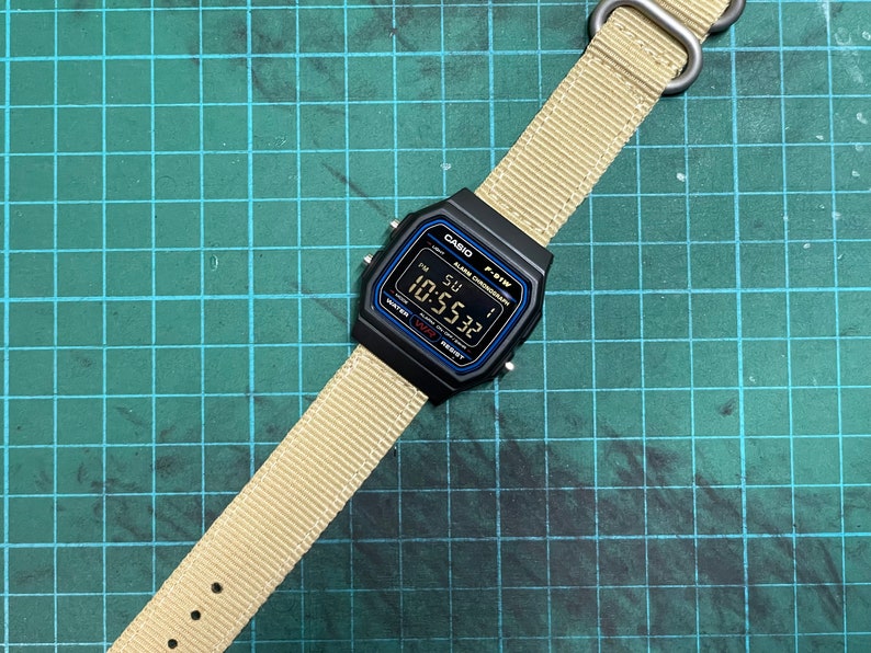 Peut inclure: Une montre num&eacute;rique noire Casio F-91W avec un bracelet en nylon beige. Le cadran de la montre affiche l'heure, la date et le jour de la semaine. La montre est sur un tapis de coupe vert.