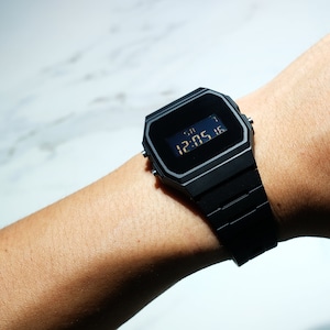 Op de afbeelding: Een zwart digitaal polshorloge met een zwarte band. Het horloge laat de tijd 12:05:16 zien.