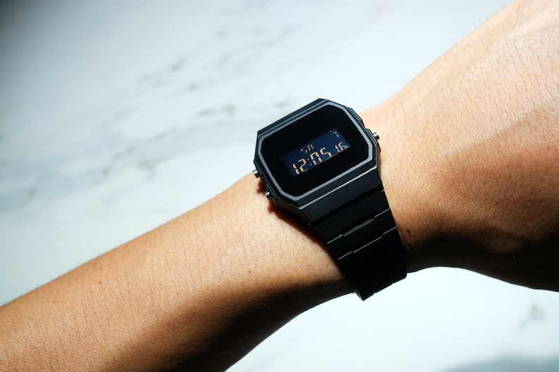 Op de afbeelding: Een zwart digitaal polshorloge met een zwarte band. Het horloge laat de tijd 12:05:16 zien.