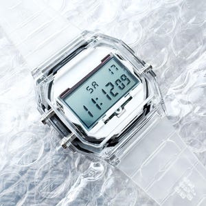 Puede incluir: Un reloj digital transparente con una esfera blanca y una correa transparente. El reloj muestra la hora y la fecha. El reloj est&aacute; hecho de pl&aacute;stico transparente y tiene una esfera cuadrada. El reloj est&aacute; sobre una superficie de pl&aacute;stico de burbujas.