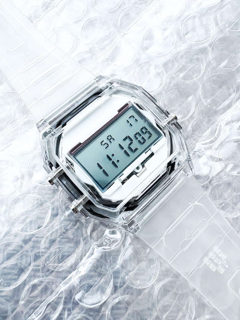 Puede incluir: Un reloj digital transparente con una esfera blanca y una correa transparente. El reloj muestra la hora y la fecha. El reloj est&aacute; hecho de pl&aacute;stico transparente y tiene una esfera cuadrada. El reloj est&aacute; sobre una superficie de pl&aacute;stico de burbujas.