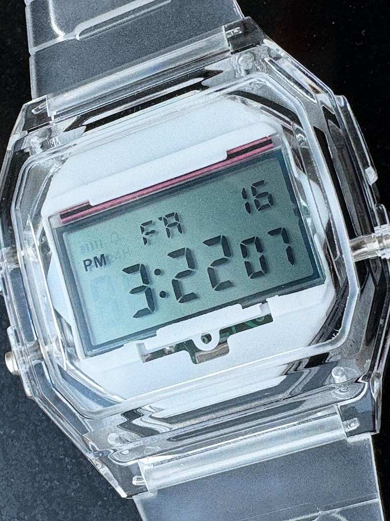 Puede incluir: Un reloj digital transparente con una esfera blanca y una pantalla rectangular. El reloj muestra la hora 3:22:07 PM del viernes 16. El reloj tiene una correa transparente y una caja octogonal transparente.