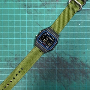 Peut inclure: Une montre num&eacute;rique noire Casio F-91W avec un bracelet en nylon vert. Le cadran de la montre affiche l'heure 1:10:09 et le jour de la semaine SU.