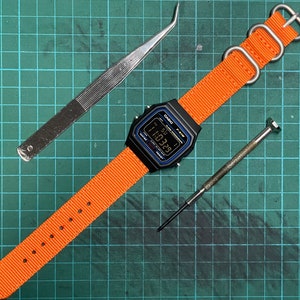 Peut inclure: Une montre num&eacute;rique noire avec un bracelet en nylon orange et une boucle argent&eacute;e. Le cadran de la montre affiche l'heure 11:39. La montre est sur un tapis de coupe vert avec une pince &agrave; &eacute;piler et un petit tournevis.
