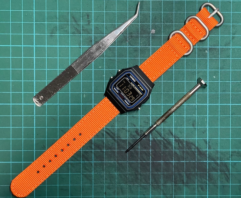 Peut inclure: Une montre num&eacute;rique noire avec un bracelet en nylon orange et une boucle argent&eacute;e. Le cadran de la montre affiche l'heure 11:39. La montre est sur un tapis de coupe vert avec une pince &agrave; &eacute;piler et un petit tournevis.