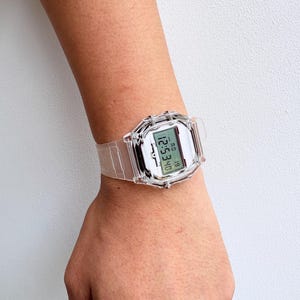 Puede incluir: Un reloj digital transparente con una esfera rectangular y una correa a juego. El reloj muestra la hora y la fecha en formato digital. El reloj se lleva en la mu&ntilde;eca.
