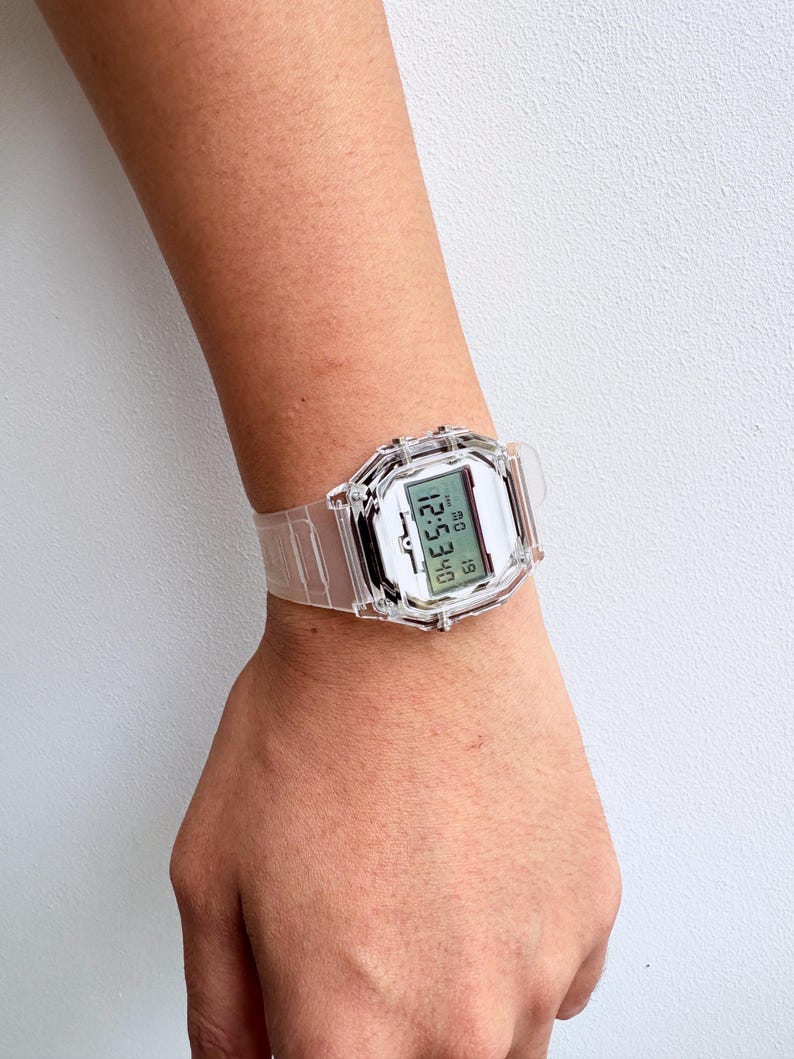 Puede incluir: Un reloj digital transparente con una esfera rectangular y una correa a juego. El reloj muestra la hora y la fecha en formato digital. El reloj se lleva en la mu&ntilde;eca.