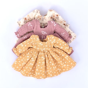 Peut inclure: Trois robes de poup&eacute;e &agrave; imprim&eacute; floral en jaune, rose et cr&egrave;me. Les robes ont des manches longues et une jupe pliss&eacute;e.