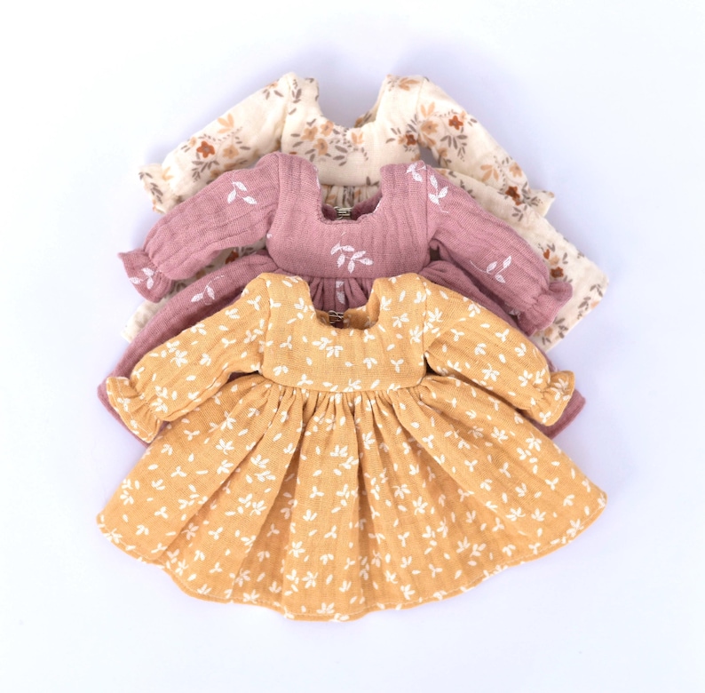 Peut inclure: Trois robes de poup&eacute;e &agrave; imprim&eacute; floral en jaune, rose et cr&egrave;me. Les robes ont des manches longues et une jupe pliss&eacute;e.