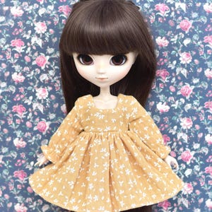 Peut inclure: Une poup&eacute;e portant une robe jaune &agrave; imprim&eacute; floral blanc et de longs cheveux bruns. La poup&eacute;e a de grands yeux bleus et un teint clair.