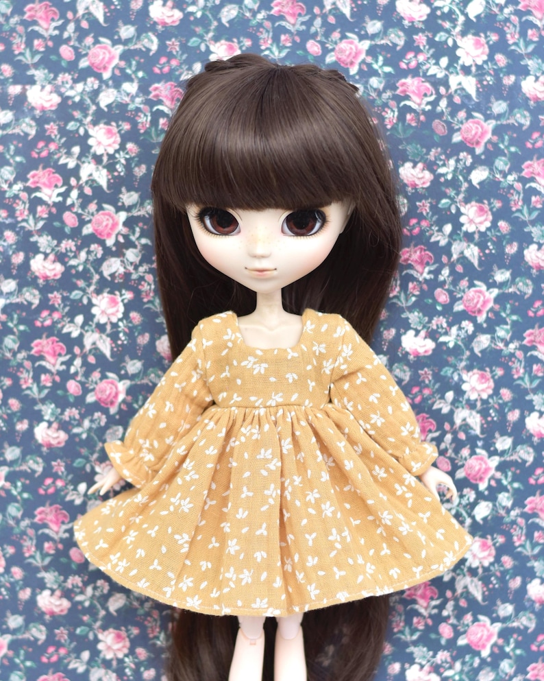 Peut inclure: Une poup&eacute;e portant une robe jaune &agrave; imprim&eacute; floral blanc et de longs cheveux bruns. La poup&eacute;e a de grands yeux bleus et un teint clair.