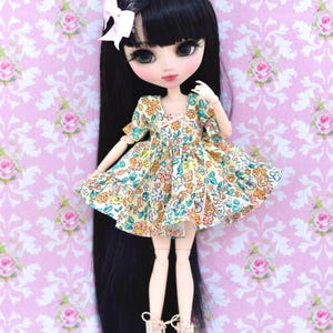 Può includere: Una bambola con lunghi capelli neri, che indossa un vestito a stampa floreale e pattini rosa. La bambola ha grandi occhi e un fiocco rosa nei capelli.